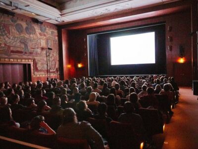 Foto (von honten): In einem Kinosaal blicken die Zuschauer auf die noch weiße Leinwand